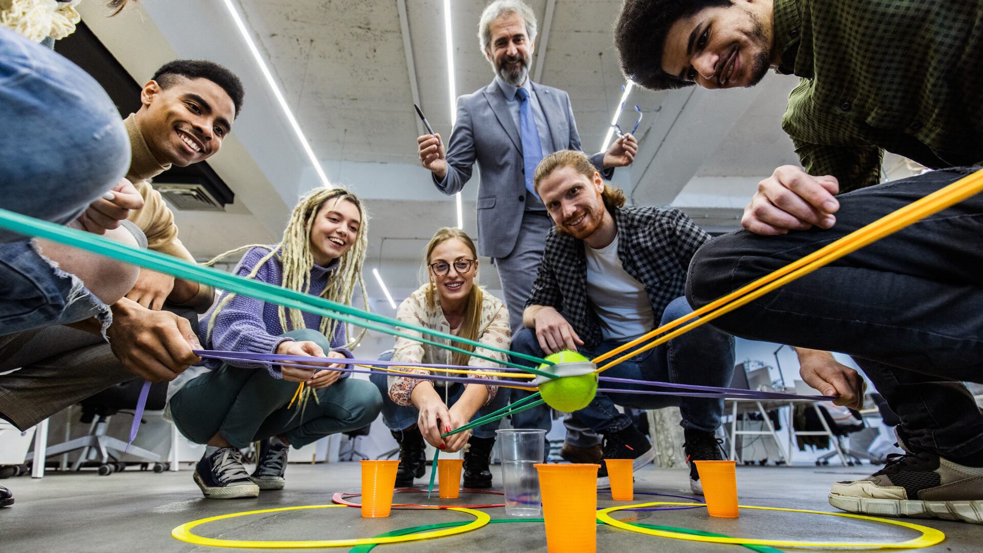 team building activities - diverse group of office workers crouching in a circle each hold rubber bands that hold a ball over a cup
