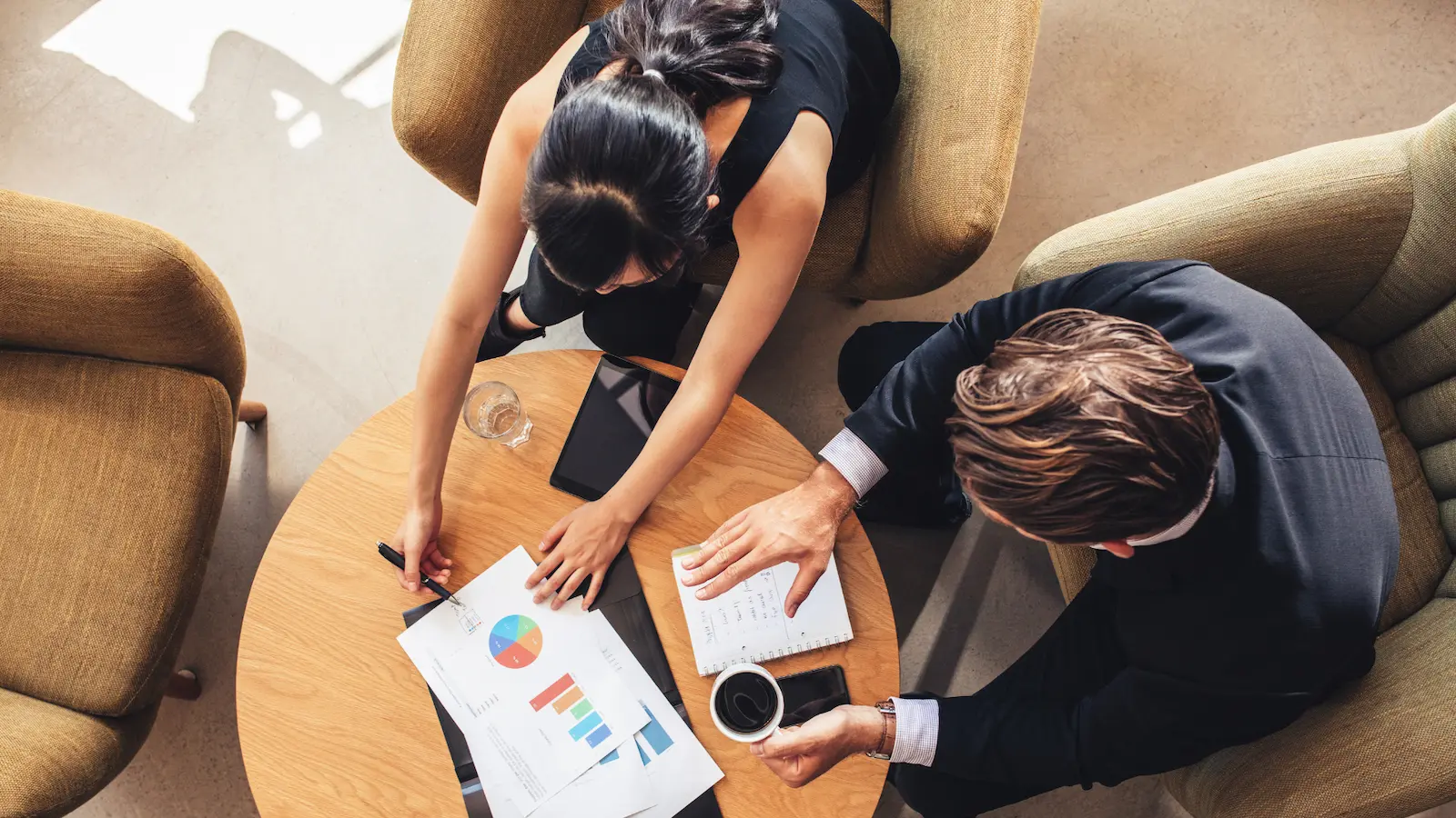 administrative assistant strategic partnership - overhead photo looking down at man and woman at a table. she is showing him some graphs, and he is has his hand flexed as if he's actively listening