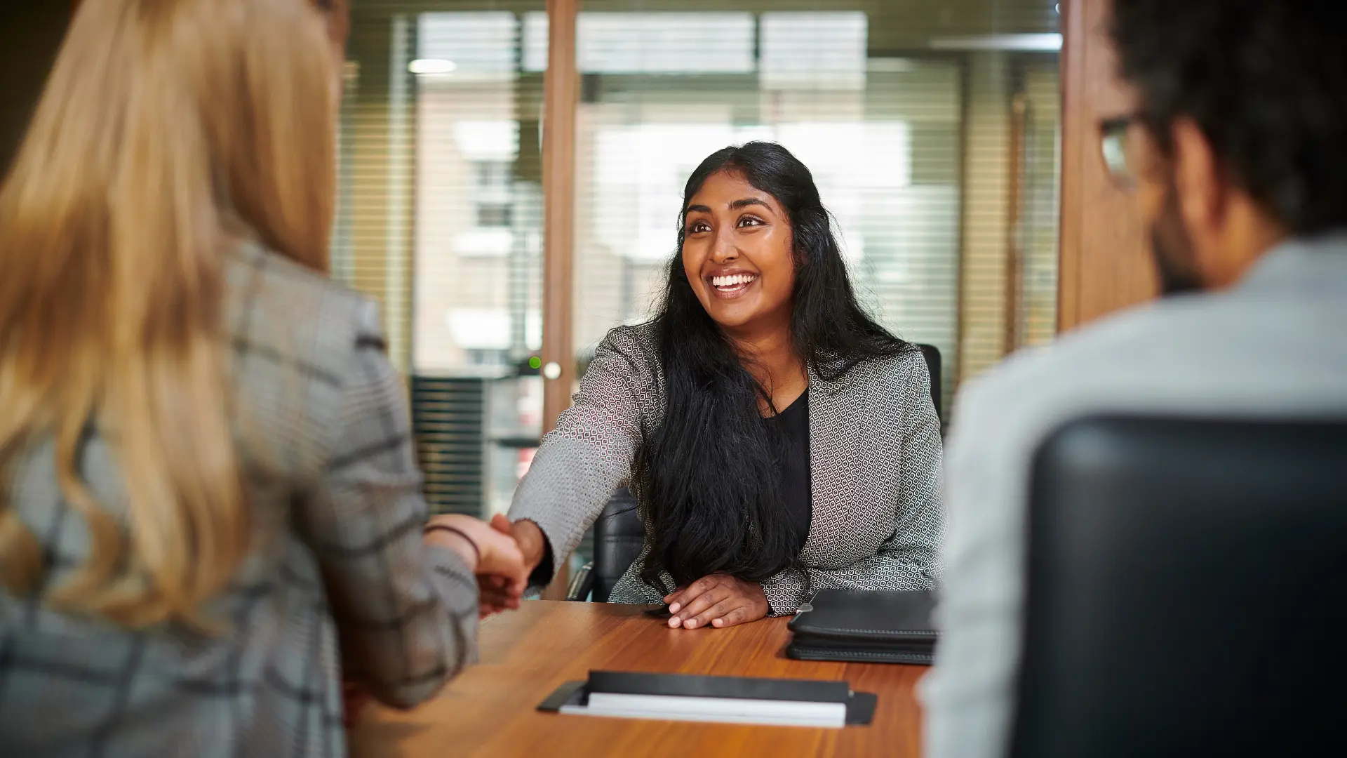 administrative and executive assistant careers blog image shows smiling indian woman in business suit shaking hands across desk with blonde woman as brunette man in glasses looks on