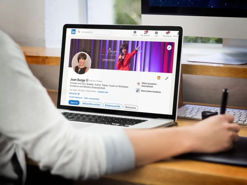 Image for LinkedIn profile tips for assistants shows arm and hand of someone sitting at a computer with a LinkedIn profile showing on the screen.