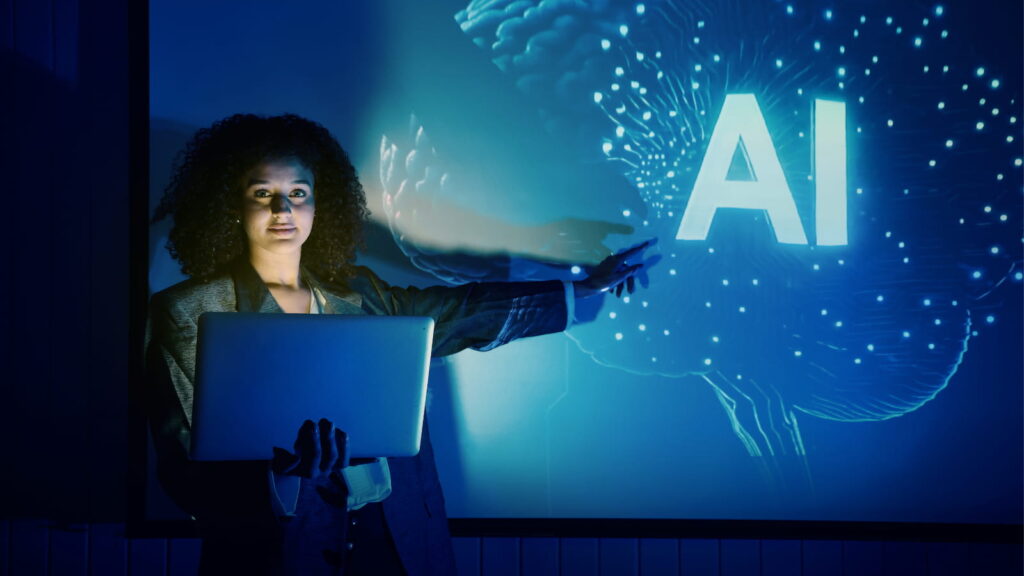 AI training for administrative professionals blog image shows a woman with a laptop in a darkened room pointing to a projection of a brain with the letters "AI"
