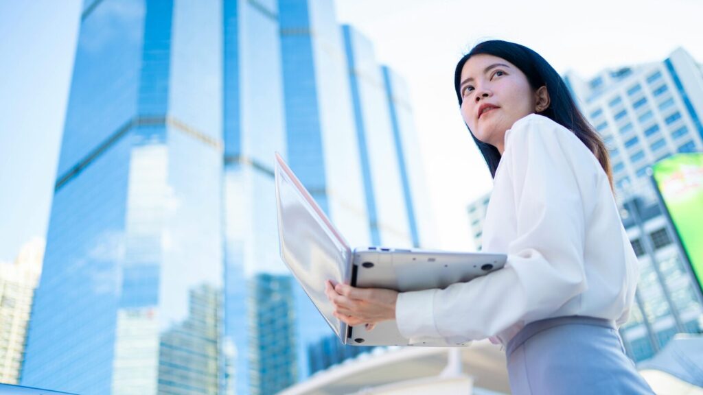 Image for Whats holding assistants back in their careers blog shows woman holding laptop with skyscrapers in background looking backward with regret